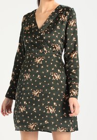 Robe florale verte avec un design enveloppant, manches longues et petites fleurs brunes et beiges. Fabriquée en tissu lisse avec une texture douce.