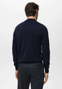 Een man die met zijn rug naar de kijker staat, gekleed in een marineblauwe trui met lange mouwen en donkergrijze broek, tegen een effen lichte achtergrond.