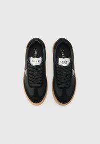 Svart sneaker med en glatt og semsket tekstur, med svarte snører, en hvit tunge med branding, og en lys brun gummisåle.