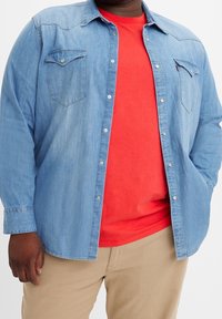 Mężczyzna w otwartej niebieskiej koszuli dżinsowej na czerwonej koszulce i beżowych spodniach, z jedną ręką w kieszeni.