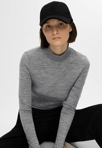 Grå langermet skjorte med høy hals, teksturert stoff, kombinert med en svart caps med buet skygge, sittende mot en nøytral bakgrunn.