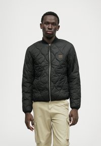 Schwarze gesteppte Bomberjacke mit Reißverschluss vorne, geripptem Kragen und einem kleinen braunen Logopatch. Getragen mit hellen Cargo-Hosen.