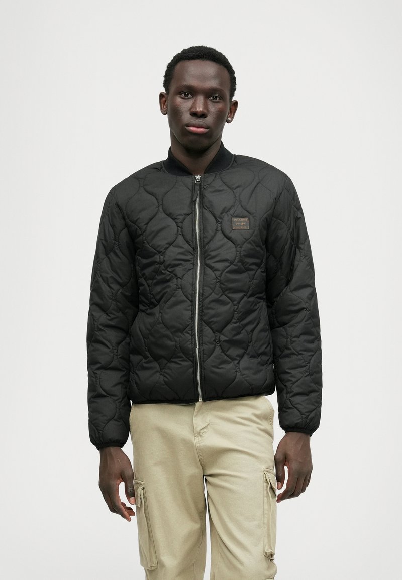 Schwarze gesteppte Bomberjacke mit Reißverschluss vorne, geripptem Kragen und einem kleinen braunen Logopatch. Getragen mit hellen Cargo-Hosen.