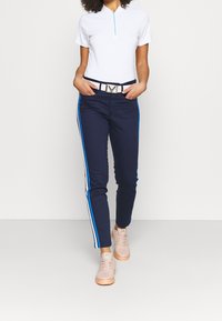 Vit kortärmad polo med blå dragkedja, marinblå byxor med blå sidostrikar och ljusrosa sneakers. Silverfärgad bälte med framträdande spännen.