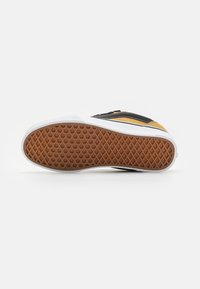 Adidași galbeni și negri cu talpă albă, prezentați din lateral, evidențiind talpa din cauciuc cu model în vafe maronii pe un fundal alb.