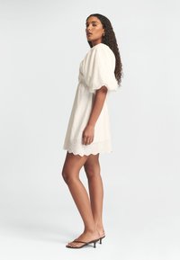 Weißes Kleid mit Puffärmeln, gewelltem Saum und Verstärkungen. Der Stoff wirkt leicht, und die Silhouette ist lässig und oberhalb des Knies.