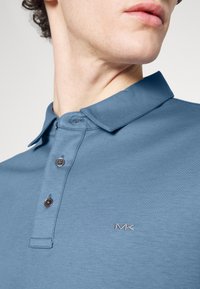 Mėlynos polo marškiniai su struktūruotu apykakle, trimis mygtukais ir išsiuvinėtu "MK" logotipu kairėje krūtinės pusėje. Lygi audinio tekstūra.