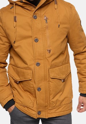 Veste à capuche marron avec fermetures à boutons et à glissière, poches à rabat avant et cordons tressés, portée avec un pantalon gris.