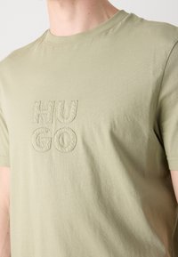 T-shirt vert olive clair à manches courtes avec lettrage "HUGO" cousu sur la poitrine, porté par une personne.