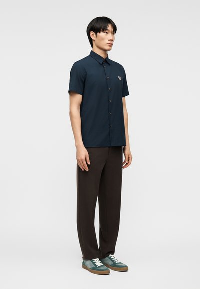 Άνδρας που φοράει σκούρο μπλε κοντομάνικο πουκάμισο με κουμπιά, σκούρο καφέ παντελόνι και τιρκουάζ αθλητικά παπούτσια, στέκεται μπροστά σε ανοιχτό γκρι φόντο.