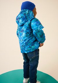 Blå mönstrad dunjacka med abstrakta mönster och färgglada dinosaurie-grafiker, i kombination med mörka jeans och vita sneakers.