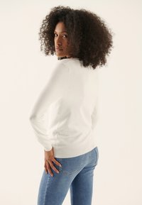 White, long-sleeve sweater with a round neck and ribbed cuffs, paired with light blue jeans. Model has curly hair and a side profile.