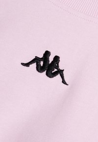 El sudadera de color rosa claro tiene un logo bordado en negro que representa figuras estilizadas sentadas. La tela muestra una textura suave y lisa.