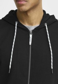 Haut à capuche zippé noir avec intérieur gris, présentant des cordons de serrage blancs avec des motifs noirs et un tirette de fermeture éclair argentée. Tissu doux et texturé.