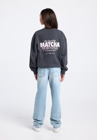 Oversized grijze trui met roze grafische tekst op de achterkant, gecombineerd met lichtblauwe wijde jeans en witte sneakers. Gladde textuur.