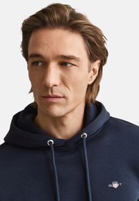 Marineblauwe hoodie met een trekkoord en een klein logo op de borst. Zachte stof en een comfortabele, losse pasvorm, met een ontwerp van een kangoeroezak.