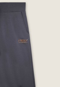 Joggers grigi scuri realizzati in tessuto morbido, con una tasca laterale e un piccolo logo metallico di colore rame sul davanti.