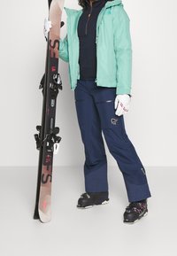 Die Skiausrüstung umfasst eine mintgrüne wasserdichte Jacke, marineblaue Skihosen mit Reißverschlüssen, weiße Handschuhe und einen schwarzen Ski mit roten Akzenten.