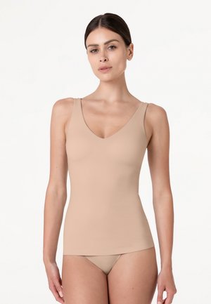 Donna con capelli scuri che indossa un top beige aderente senza maniche e biancheria intima abbinata su uno sfondo bianco uniforme.