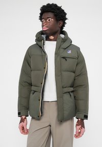 Groene pufferjack met een capuchon, met een gewatteerd ontwerp, ritszakken en een logo aan de voorkant. Gecombineerd met een wit shirt en beige broek.