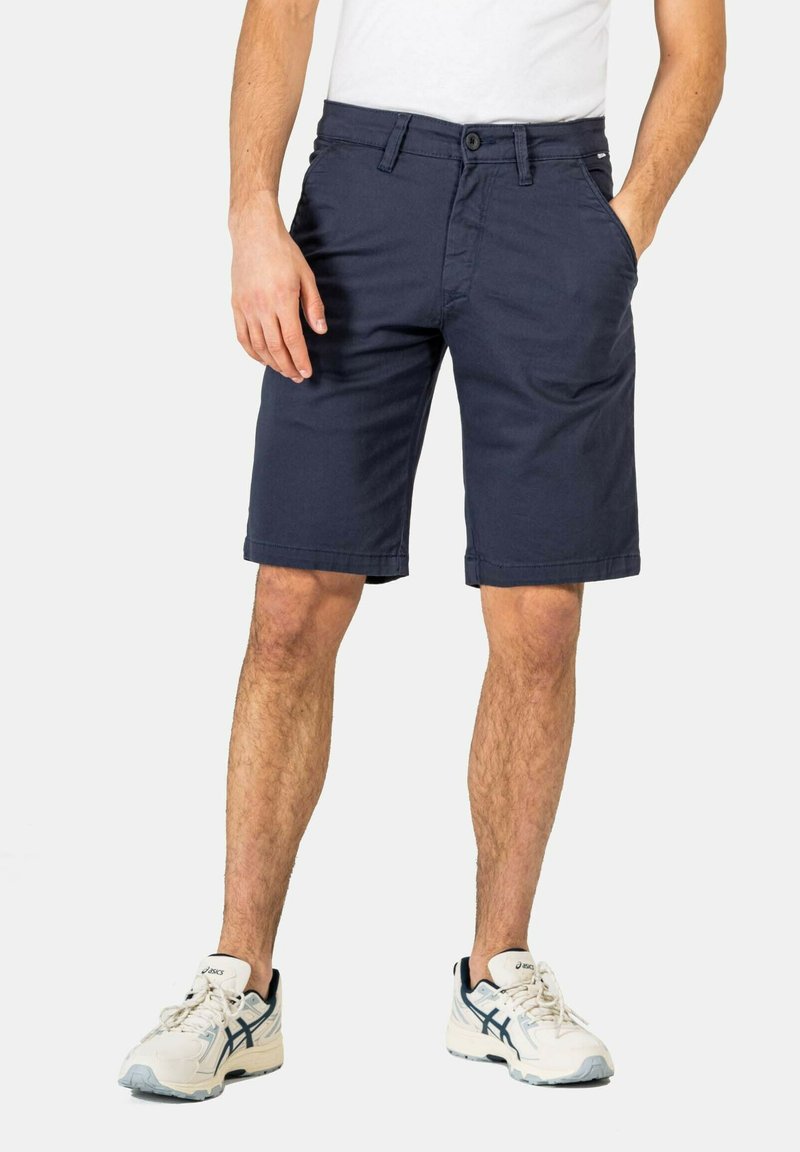 Navy blue cotton shorts with a straight cut, front pockets, and a button closure. Worn with a white t-shirt and light-colored sneakers.