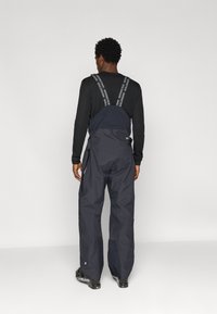 Fekete vízálló munkanadrág, állítható vállpántokkal, laza szabással és oldalsó zsebekkel. Az anyag tartósnak tűnik, matt felülettel.