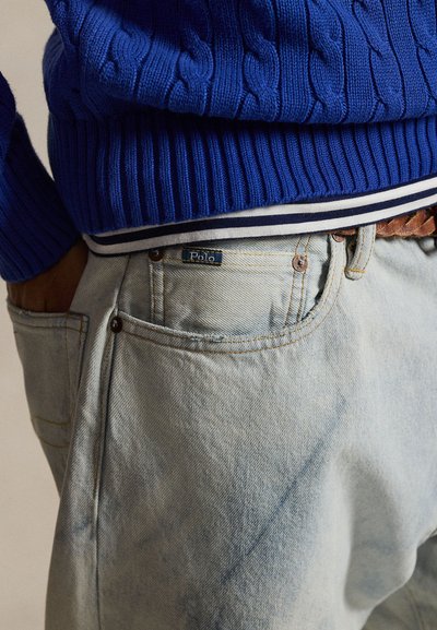 Personne portant un jean délavé clair avec une étiquette "Polo", une ceinture en cuir tressé, un pull à torsades bleu et une chemise rayée en dessous.