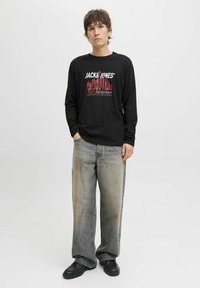 Joven de pie con las manos en los bolsillos, vistiendo una camisa de manga larga negra de "Jack & Jones", jeans anchos y desgastados, y zapatos negros sobre fondo blanco.