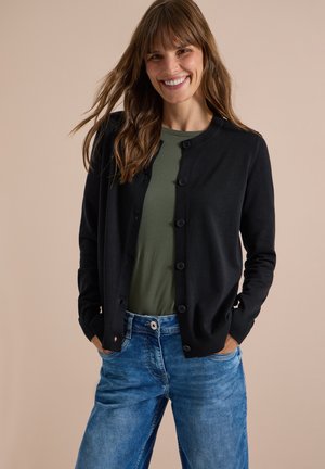 Femme souriante aux longs cheveux bruns porte un cardigan noir boutonné, une chemise verte et un jean bleu, debout les mains dans les poches devant un fond beige.