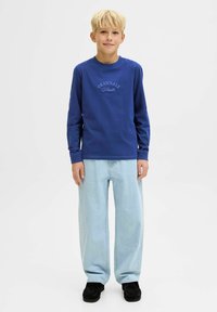 Sudadera de manga larga azul con el texto "ORIGINALS" en letras en relieve, combinada con jeans de pierna ancha de color azul claro y zapatos negros.