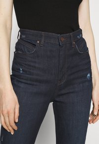 Mörk tvättade denimjeans med hög midja, fem fickor och lätt slitningar på låren. Kopparfärgad hårdvara och kontrasterande sömmar.