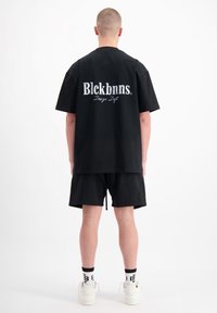 Zwarte katoenen oversized t-shirt, korte mouwen, met wit geborduurd "Blckbnns" logo en "Design Dept" op de achterkant. Draag het met zwarte shorts.