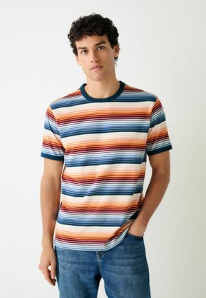 Junger Mann steht mit der Hand in der Tasche, trägt ein mehrfarbig gestreiftes T-Shirt mit horizontalen Streifen und blaue Jeans vor einem einfarbigen Hintergrund.