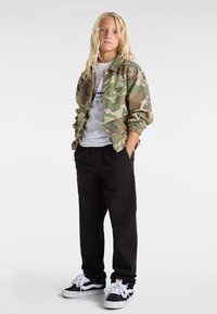 Kamikaze-jakke i camouflagemønster i grønne og brune nuancer, parret med en hvid t-shirt og sorte bukser; sorte og hvide sneakers med snørebånd.