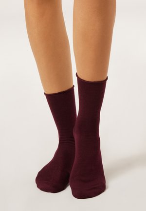 Jambes portant des chaussettes de longueur mi-mollet de couleur bordeaux foncé, sur un fond clair uni.