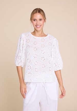 Smilende kvinne iført en hvit, blomstrete bluse med hullmønster og puffede ermer, og hvite bukser med snor, mot en beige bakgrunn.