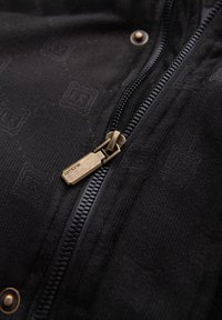 Svart tyg med ett texturerat mönster, med en framträdande dragkedjeskötsel i mässing och en tryckknapp för stängning.