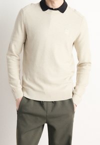 Suéter de punto beige texturizado con cuello negro, con dobladillo y puños acanalados, usado sobre pantalones verde olivo oscuro.