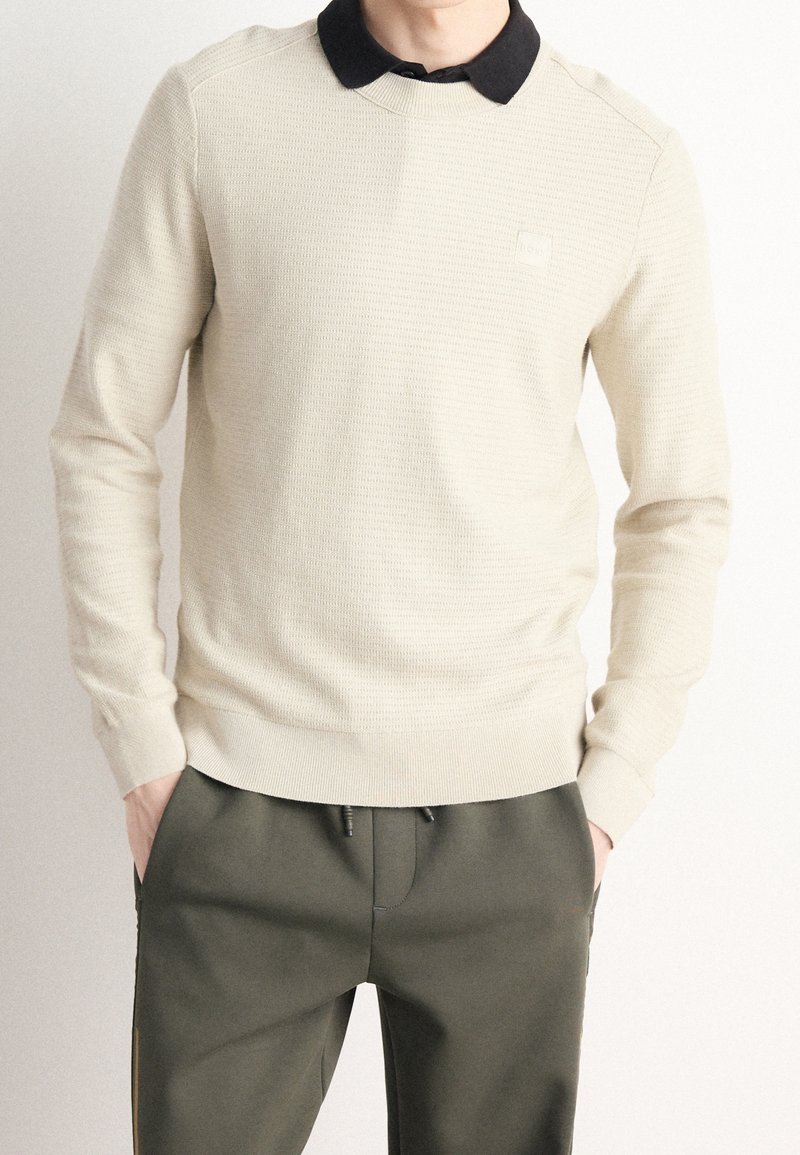 Suéter de punto beige texturizado con cuello negro, con dobladillo y puños acanalados, usado sobre pantalones verde olivo oscuro.