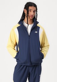 Veste de sport bleu marine et jaune avec fermeture éclair devant, deux poches latérales et taille élastiquée. Présente un design à panneaux contrastés et un logo sur la poitrine.