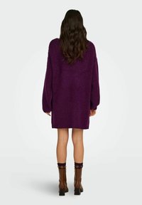 Oversizeowy fioletowy sweter dziany z opuszczanymi ramionami, teksturowaną tkaniną i ściągaczami na rękawach; zestawiony z brązowymi botkami na obcasie i wzorzystymi skarpetkami.