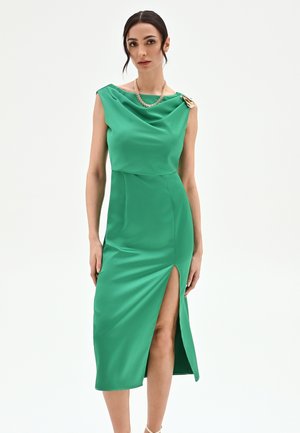 Femme vêtue d'une robe émeraude sans manches avec une fente haute sur la cuisse, portant un collier chaîne en or et une fermeture décorative sur une épaule, sur un fond uni.