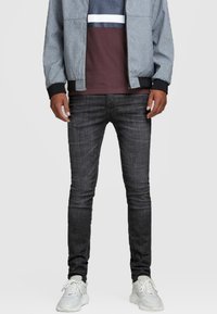 Jeans skinnies gris oscuro con una textura suave, combinados con una camisa a bloques de color burdeos y gris y una chaqueta bomber gris. Zapatillas blancas completan el atuendo.
