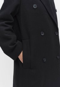 Svart dubbelknäppt kappa med två knappar, sidofickor och ett texturerat tyg. Har en klassisk kragdesign och en avslappnad passform.