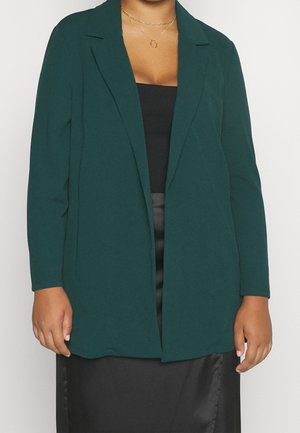 Kvinde iført mørkegrøn åben blazer over sort top og sort nederdel, med to delikate gyldne halskæder.