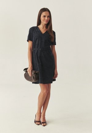 Sieviete, tērpusies tumši zilā kleitā ar īsām piedurknēm un jostu, turot tumši brūnu rokassomu un valkājot atvērtu purngalu augstpapēžu kurpes uz gaiša fona.