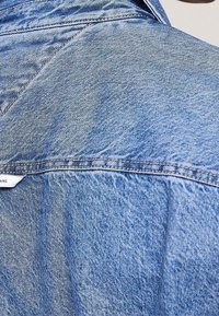 Close-up of back shoulder area of light blue denim jacket with visible stitching and a partial white fabric label.