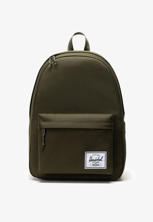 Olijfgroene stoffen rugzak met een ronde vorm, een voorvak met rits en een wit labelpatch met zwarte tekst. Glad van structuur.