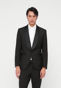 Jeune homme portant un smoking noir avec des revers en satin et une chemise blanche, se tenant contre un fond blanc uni.