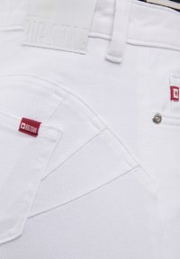 Jeans de mezclilla blanca con una superficie texturizada, acentos de logotipo rojo y un distintivo detalle de costura en los bolsillos. Incluye una etiqueta de marca en relieve blanca.